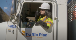 A woman wearing a hard hat, safety vest, and gloves sits in a truck labeled “Traffic Plan,” speaking into a walkie-talkie—showcasing real-world marketing in the construction industry.